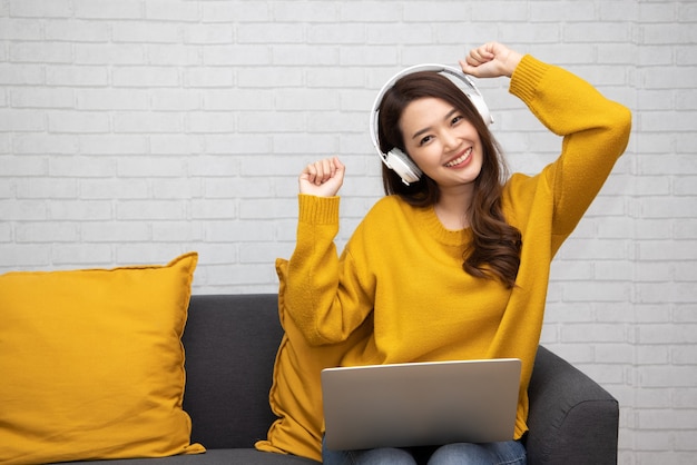 Portrait of a happy young Asian woman listening music with wireless headphones from music application in computer laptop