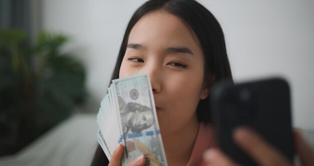 Photo portrait of happy young asian woman sitting in an excited state holding an online smartphone and dollar bills in her hand saves money