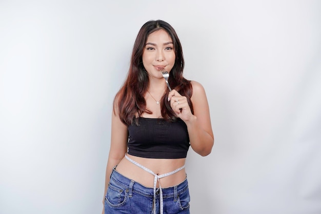 Portrait of happy young Asian woman wearing body measuring tape around waist holding spoon and fork isolated over white background