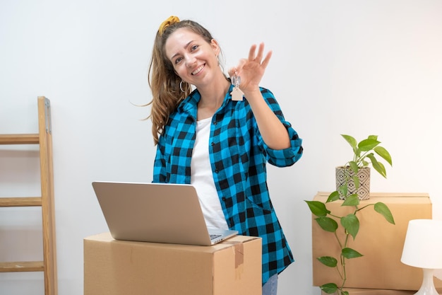 Portrait of happy young attractive woman with laptop on cardboard boxes moving in a new home.