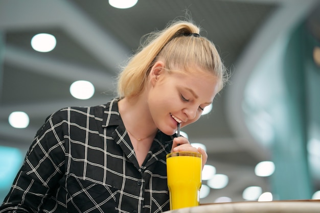 カフェでオレンジジュースを飲んで幸せな若いブロンドの女の子の肖像画。レジャーと人々のコンセプト