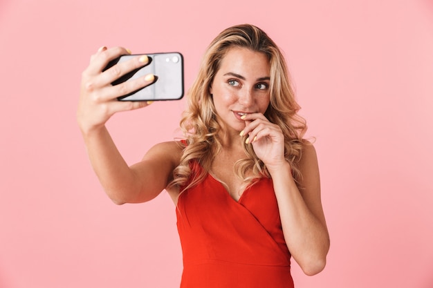 Portrait of a happy young blonde woman posing isolated over pink wall talking by mobile phone take a selfie