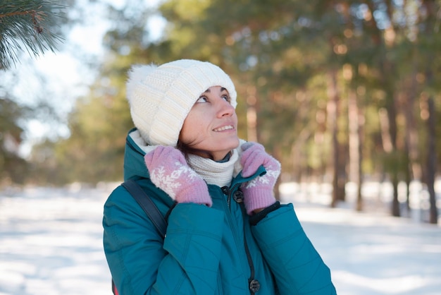 Portrait of a happy young brunette in winter clothes in a white knitted hat in the frost in the sun