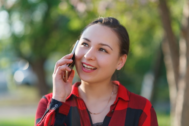 スマートフォンを手に持った、幸せな若いブルネットの女性の肖像画。女性の携帯電話で話していると笑顔します。晴れた日に撮影