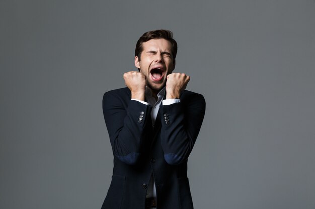 Portrait of a happy young businessman dressed in suit isolated over gray wall, celebrating success