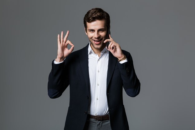 Portrait of a happy young businessman dressed in suit isolated over gray wall, talking on mobile phone, showing ok gesture