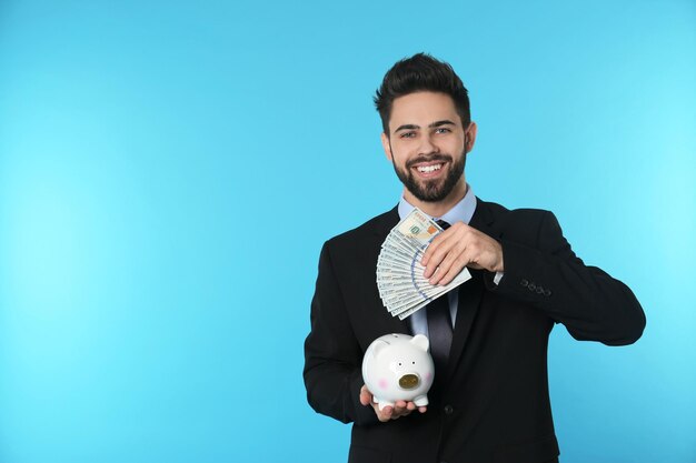 Portrait of happy young businessman with money and piggy bank on color background Space for text