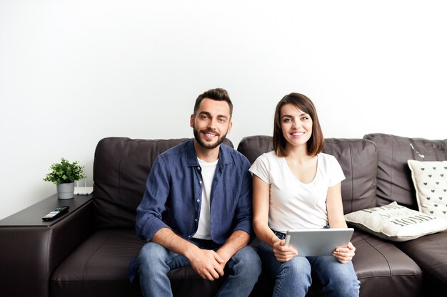 Portrait of happy young couple in casual outfits sitting on comfortable sofa at home and using digital tablet to watch movie