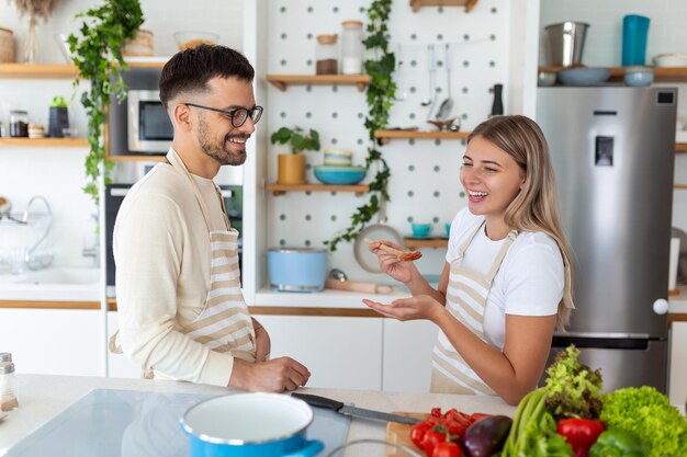 自宅のキッチンで一緒に料理をする幸せな若い夫婦のポートレート ロマンチックな魅力的な若い女性とハンサムな男性は、明るいモダンなキッチンに立ちながら、一緒に過ごす時間を楽しんでいます