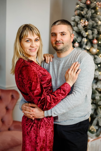 Portrait of happy young couple hugging, cuddling, celebrating winter holidays together near decorated Christmas tree in living room.