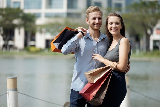 Portrait of happy young couple in love holding shopping bags when standing outdoors and smiling at c...