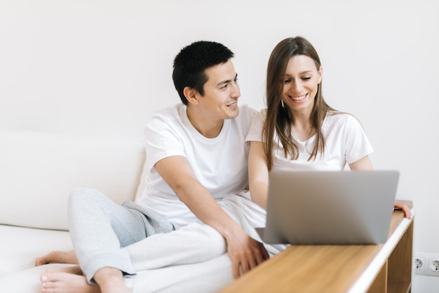 소파에 앉아서 집에서 노트북 작업을 하는 행복한 젊은 커플의 초상화. 행복한 젊은 커플이 밝은 아파트에서 노트북 작업을 하고 있습니다.
