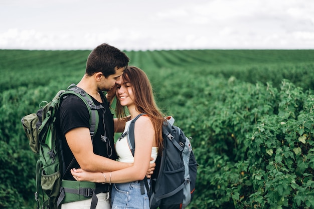 Portrait of happy young couple with backpacks on the field in spring. Man and woman walking in currant plantations