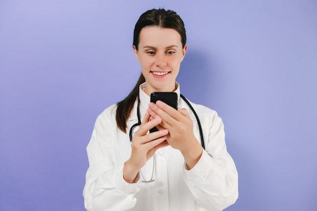 Foto ritratto di giovane dottoressa felice che tiene lo smartphone guarda lo schermo indossa lo stetoscopio e l'uniforme bianca, l'innovazione del sito web della rete di prescrizione online, in posa isolata sopra la parete di sfondo viola dello studio