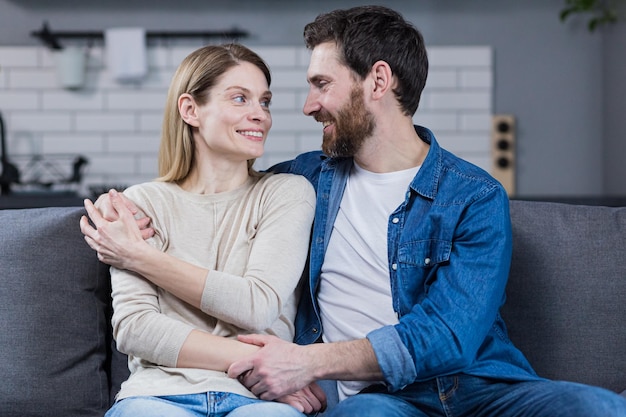 Portrait of a happy young family couple Man and woman sitting on sofa hugging waving smiling looking at camera looking at each other