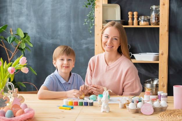 Portrait of happy young family sitting at wooden table with paint cans and preparing for Easter