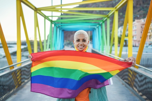 LGBTの虹色の旗を保持している幸せな若いゲイの女性の肖像画-顔に焦点を当てる