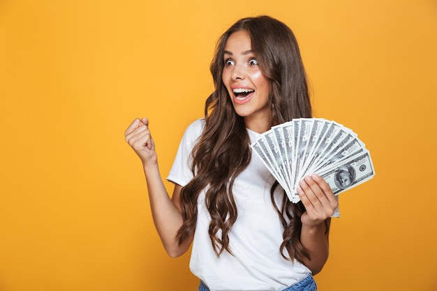 Portrait of a happy young girl with long brunette hair standing over yellow wall, holding money banknotes, looking away