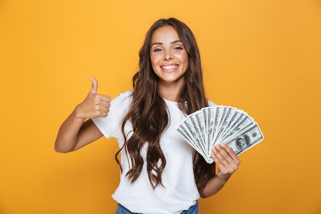 Photo portrait of a happy young girl with long brunette hair standing over yellow wall, holding money banknotes, showing thumbs up