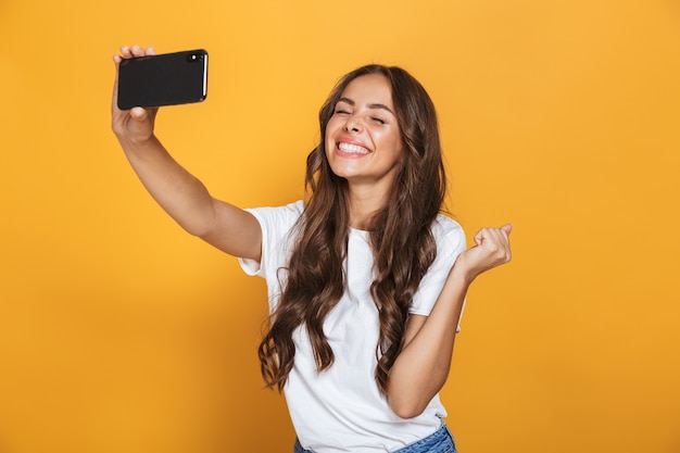 Portrait of a happy young girl with long brunette hair standing over yellow wall, taking a selfie