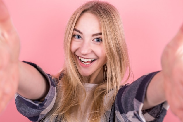 Portrait of a happy young girl with straight hair, in a shirt, taking a selfie on a pink wall