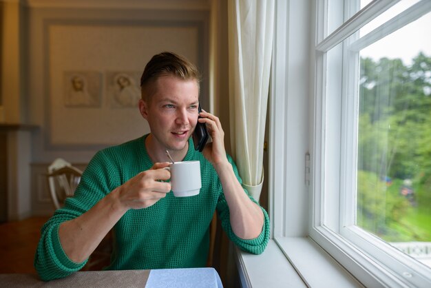 電話を使用して、自宅でコーヒーを飲む幸せな若いハンサムな男の肖像画