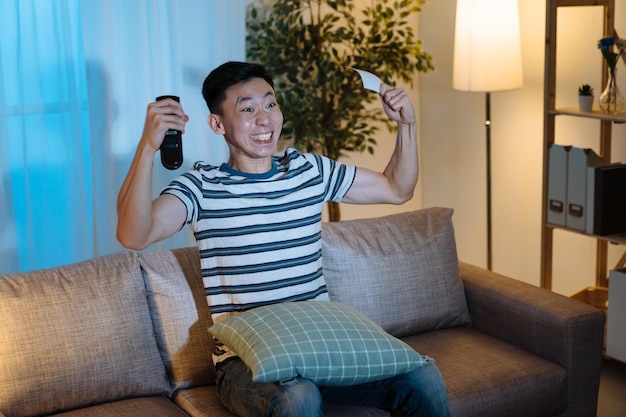 Portrait happy young japanese man with pumping fists ecstatic\
celebrates success in dark living room in apartment at night.\
cheerful guy sitting on couch hand hold remote control and lottery\
ticket