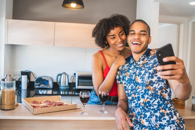 Portrait of happy young latin couple taking a selfie with mobile phone while having dinner at new home. Lifestyle and relationship concept.