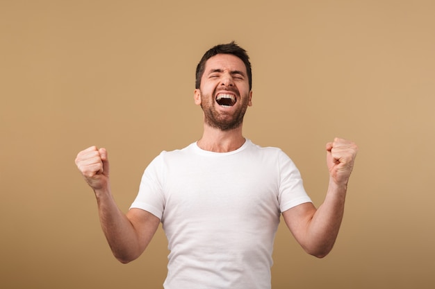 Portrait of a happy young man casually dressed standing isolated over beige, celebrating success