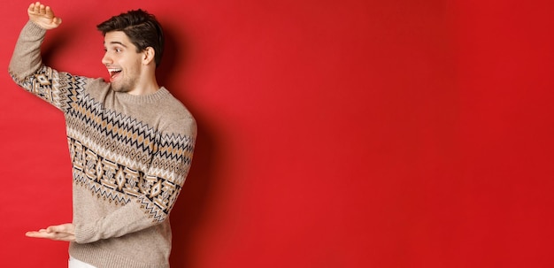 Portrait of happy young man in christmas sweater, showing large present, smiling and looking amazed at cool gift, standing over red background