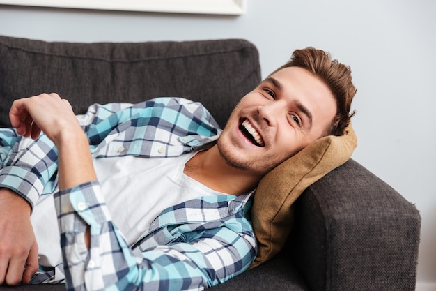 Portrait of happy young man dressed in shirt in a cage print lies on sofa at home and looking aside.