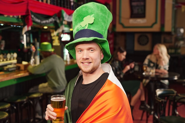 Foto ritratto di un giovane felice con un cappello verde che beve birra e celebra il giorno di san patrizio al pub