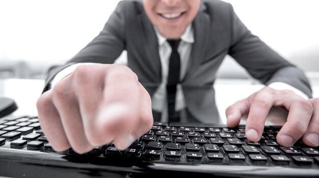 Portrait of a happy young man pointing at camera while sitting with laptop computer isolated in office