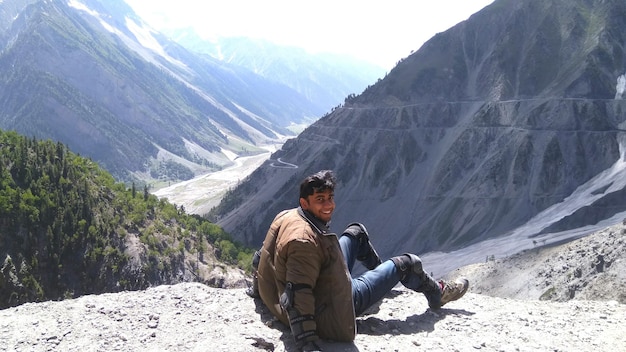 Foto ritratto di un giovane felice seduto su una scogliera contro le montagne