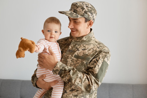 Ritratto di felice giovane soldato che indossa un'uniforme mimetica e berretto in posa con la sua simpatica bambina neonato che tiene un peluche guardando la fotocamera nelle mani del padre