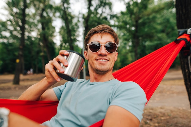 彼の手に温かい飲み物のカップとハンモックに横たわっているサングラスで幸せな若い男の肖像画