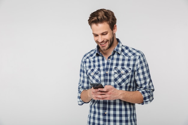 Ritratto di un giovane felice che indossa una camicia a quadri sorridente e usa il cellulare isolato su un muro bianco