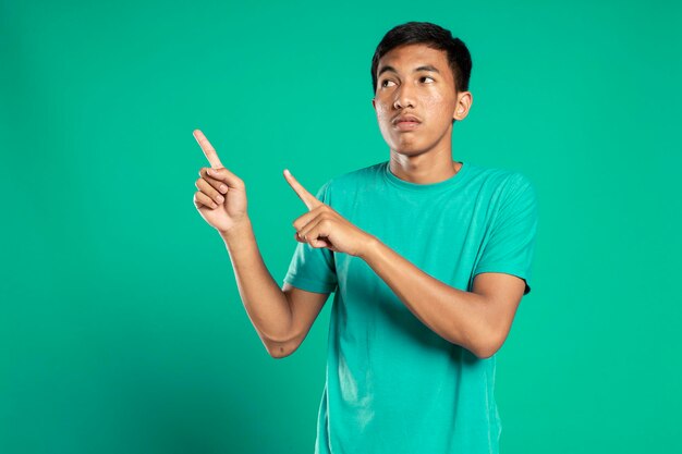 Portrait of a happy young man in white t-shirt pointing fingers away at copy space isolated over green background