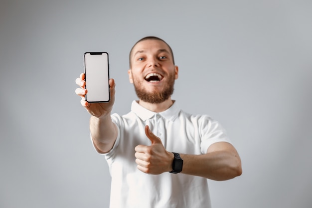 Portrait of a happy young man in a white t-shirt showing a phone screen on gray.