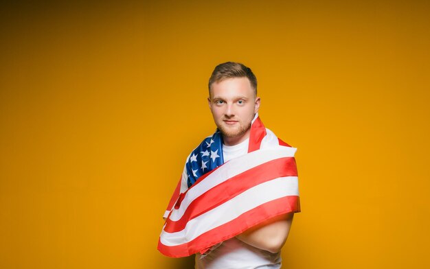 Portrait of a happy young man with a beard in casual clothes holding the us flag on a yellow