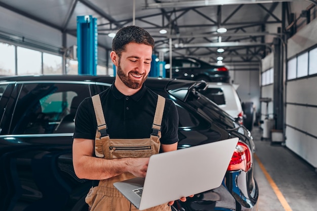 バックグラウンドでラップトップ車と修理サービスステーションで幸せな若いメカニックの肖像画