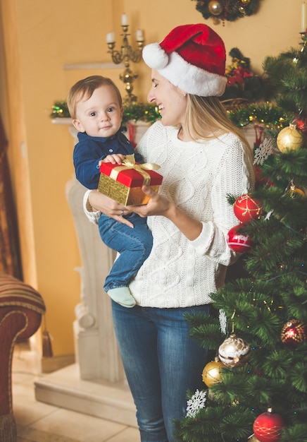 クリスマスツリーでサンタの帽子で彼女の男の子を抱きしめる幸せな若い母親の肖像画