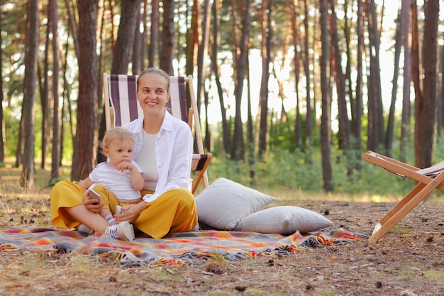 Portrait of happy young mother sitting on blanket in forest with her infant baby enjoying resting outdoor and spending time with her little kid mommy with child and smiling