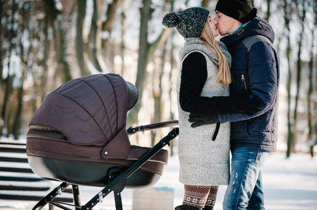 Portrait of a happy young parents stand and kissing with a stroller baby in a winter park