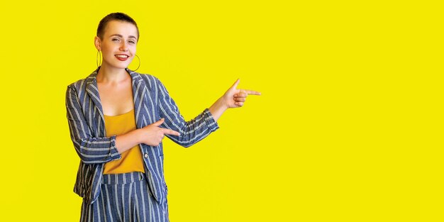 Ritratto di felice giovane capelli corti bella donna in camicia gialla e abito a righe in piedi che punta a copyspace su sfondo giallo e che guarda l'obbiettivo con sorriso aperto. girato in studio isolato.