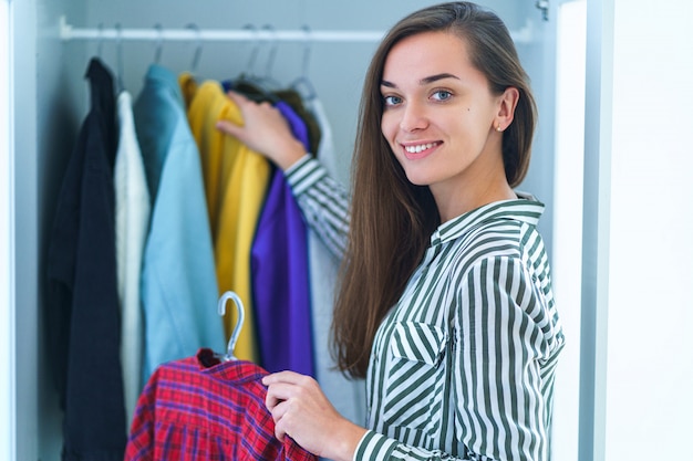 Portrait of happy young smiley brunette woman choosing outfit from wardrobe closet with stylish clothes and home stuff