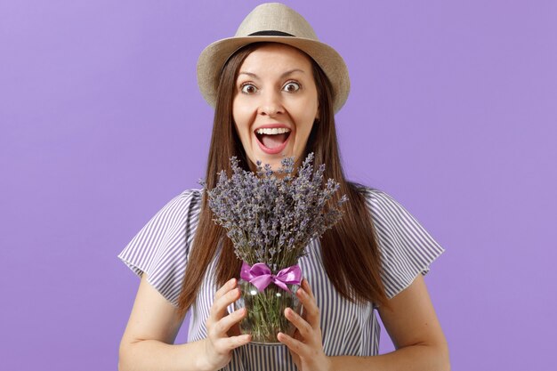 明るいトレンドの紫色の背景に分離された美しい紫色のラベンダーの花の花束を保持している青いドレス麦わら帽子の幸せな若い優しい女性の肖像画。国際女性デーの休日の概念