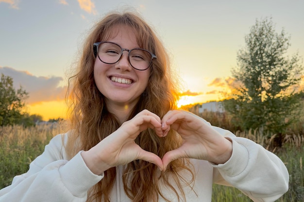 Портрет счастливой молодой женщины в очках на закате на естественном фоне, улыбающейся и смотрящей в камеру