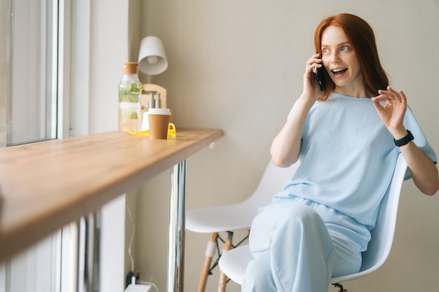 Portrait of happy young woman in good mood talking on mobile phone sitting in cozy cafe by window. 