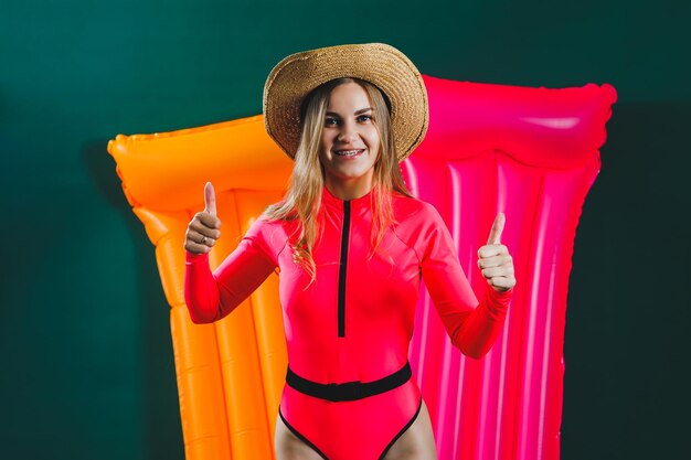 Portrait of a happy young woman in a hat dressed in a pink swimsuit smiling girl with an inflatable pink mattress isolated on a green background